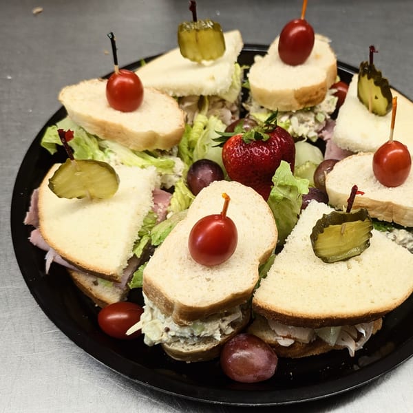 Assorted finger sandwiches with lettuce, deli fillings, pickles, cherry tomatoes, grapes, and strawberries on a black platter.