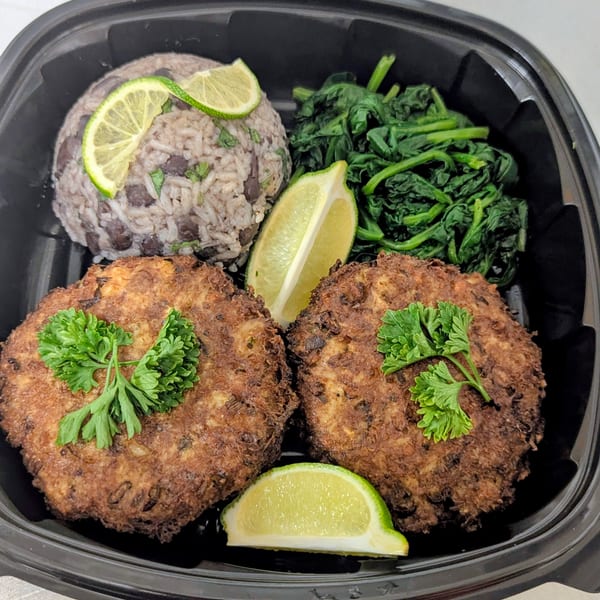 Open takeout container holding grilled meat patties, rice and beans, sautéed greens, lime wedges, and parsley garnish.